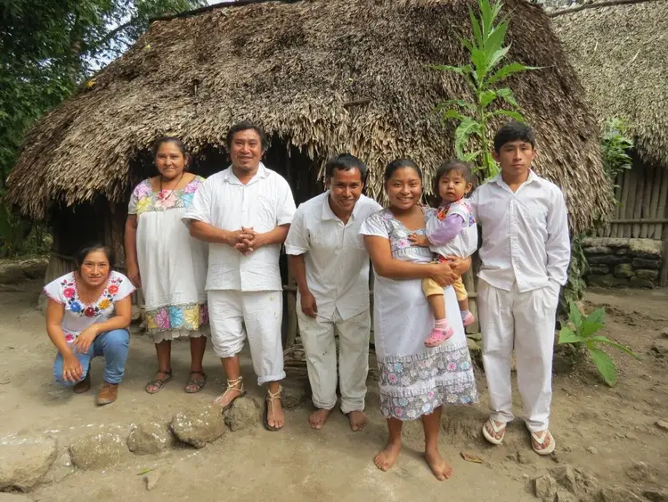 A family of seven stands together outside a thatched-roof house made of straw and wood A family of seven stands together outside a thatched-roof house made of straw and wood
