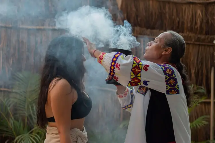Man in traditional woven white garment with colorful embroidered patterns blows smoke toward a woman during a ceremonial ritual in a thatched-roof structure Man in traditional woven white garment with colorful embroidered patterns blows smoke toward a woman during a ceremonial ritual in a thatched-roof structure
