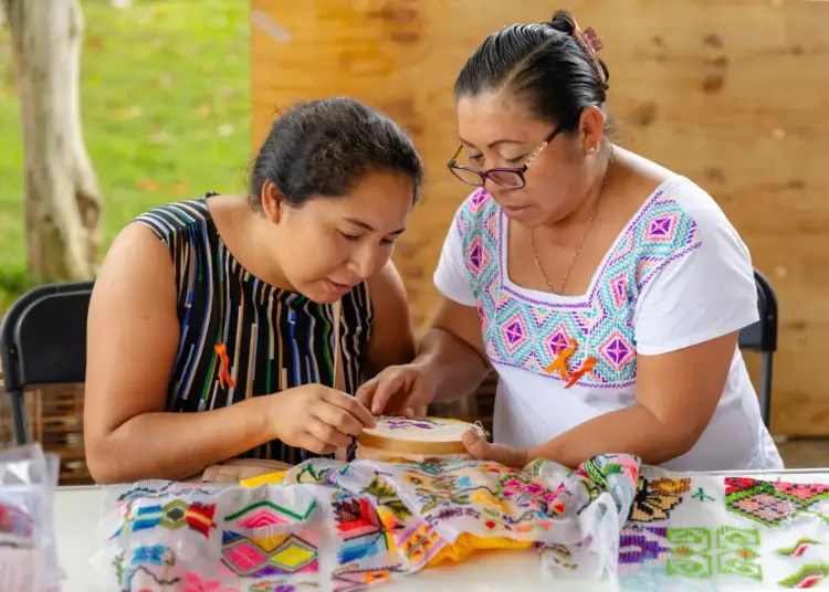 Two women work together on colorful embroidered textile with traditional geometric patterns at a wooden table outdoors Two women work together on colorful embroidered textile with traditional geometric patterns at a wooden table outdoors