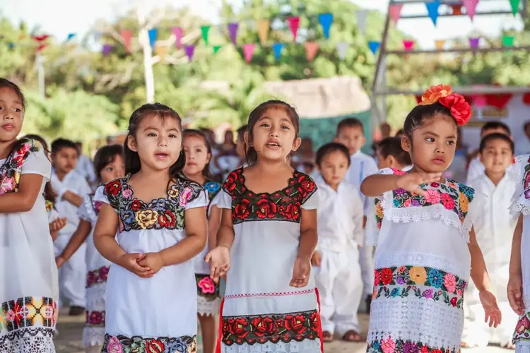 Children wearing white dresses with colorful embroidered patterns stand together outdoors under decorative bunting at what appears to be a community gathering Children wearing white dresses with colorful embroidered patterns stand together outdoors under decorative bunting at what appears to be a community gathering
