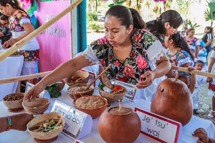 Woman in floral shirt demonstrates traditional food preparation at an outdoor community event with labeled dishes and onlookers Woman in floral shirt demonstrates traditional food preparation at an outdoor community event with labeled dishes and onlookers