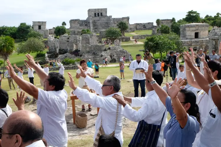 Group of people wearing white shirts with raised hands participate in a ceremony at ancient stone ruins surrounded by tourists and green vegetation Group of people wearing white shirts with raised hands participate in a ceremony at ancient stone ruins surrounded by tourists and green vegetation