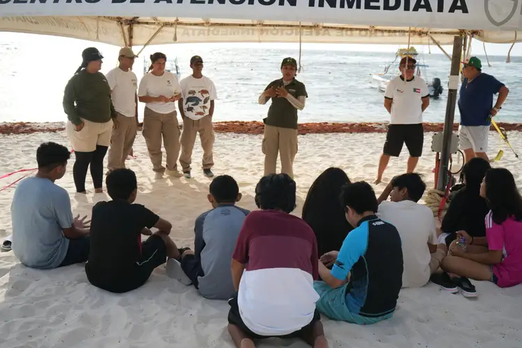 A group of people sit on sand under a tent facing uniformed officials who appear to be conducting a beach presentation near the ocean