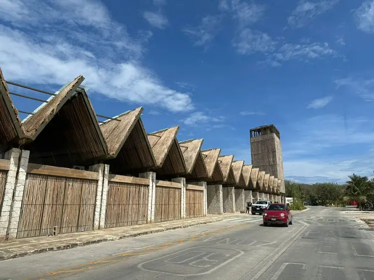 Red car drives past a long building with distinctive triangular thatched roofs and wooden slat walls along a paved street under blue sky Tulum Commercial Rental Prices Soar, Emptying Jaguar Park Shops