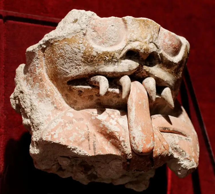 Carved stone artifact with a face featuring prominent teeth and a protruding tongue displayed against a red background