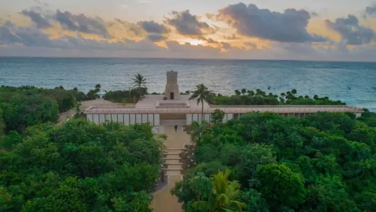 A beige stone structure with a central tower sits on a coastal peninsula surrounded by dense tropical vegetation, with turquoise ocean and cloudy sky beyond