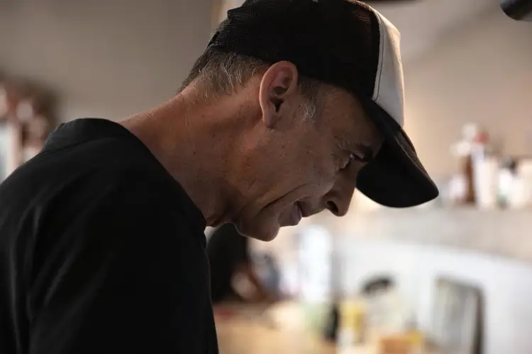 Man wearing a black cap and dark shirt looks down in an indoor kitchen setting with blurred figures and equipment in the background Man wearing a black cap and dark shirt looks down in an indoor kitchen setting with blurred figures and equipment in the background