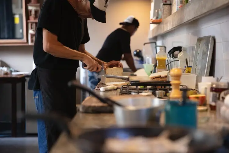 Two people prepare food in a kitchen, with one in the foreground holding a wooden spoon over a pot while another works at the counter in the background Two people prepare food in a kitchen, with one in the foreground holding a wooden spoon over a pot while another works at the counter in the background