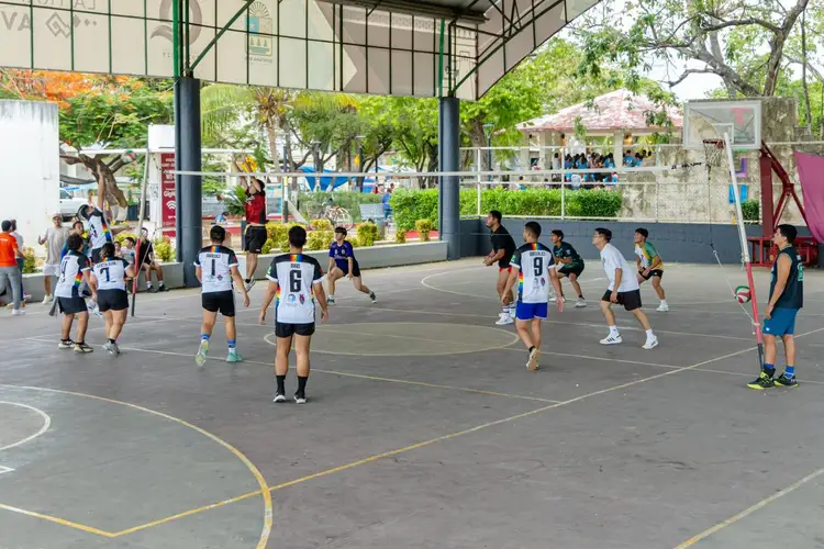 Young volleyball players in uniforms compete on an outdoor court under a covered structure with trees and buildings visible in the background Young volleyball players in uniforms compete on an outdoor court under a covered structure with trees and buildings visible in the background