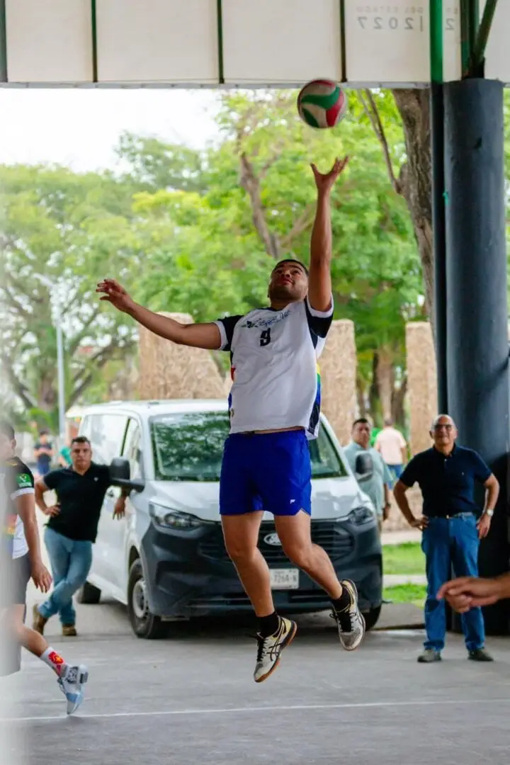 A player in a white and blue jersey jumps to catch a volleyball during an outdoor game under a covered structure, with spectators watching nearby A player in a white and blue jersey jumps to catch a volleyball during an outdoor game under a covered structure, with spectators watching nearby