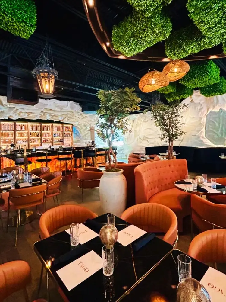 A restaurant interior features orange velvet chairs around black tables set with place settings, hanging green plant installations, copper pendant lights, and a A restaurant interior features orange velvet chairs around black tables set with place settings, hanging green plant installations, copper pendant lights, and a