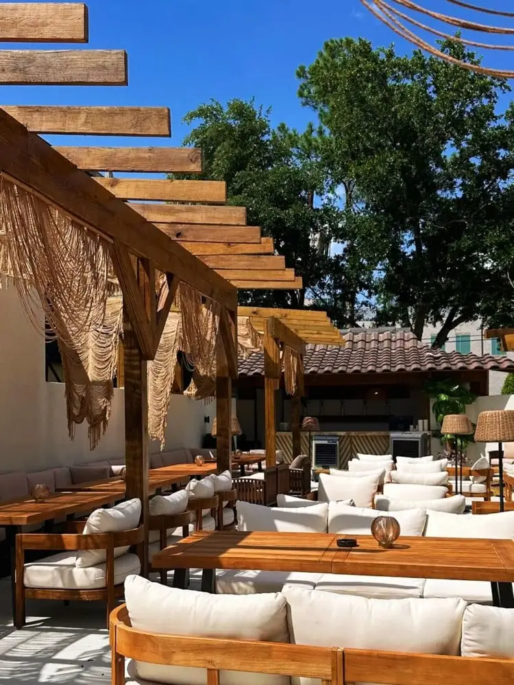 Wooden pergola with draped fabric shading lounge seating and dining tables at an outdoor patio surrounded by trees Wooden pergola with draped fabric shading lounge seating and dining tables at an outdoor patio surrounded by trees
