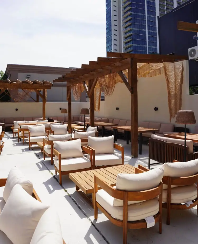 Rooftop lounge area with wooden pergolas, cream-cushioned seating, and draped fabrics overlooking tall buildings in an urban setting Rooftop lounge area with wooden pergolas, cream-cushioned seating, and draped fabrics overlooking tall buildings in an urban setting
