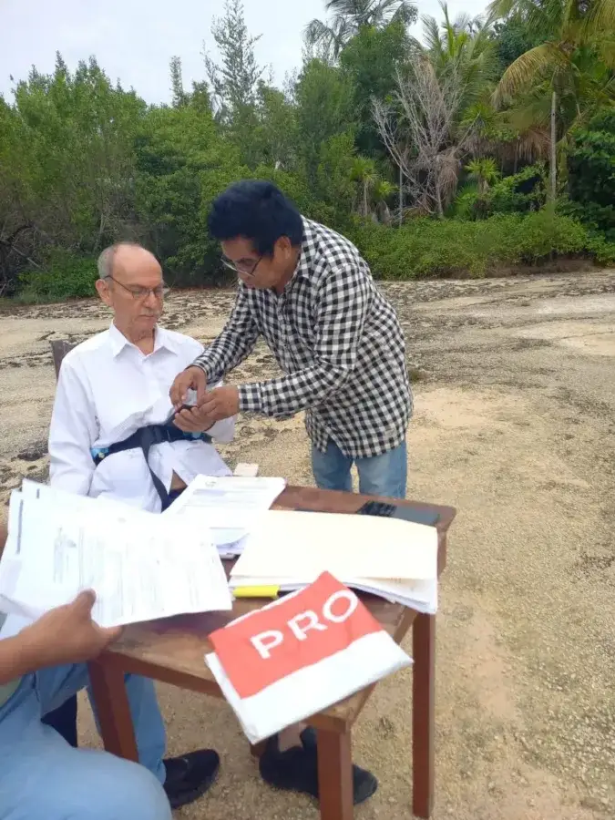 Two men review documents at a table outdoors, with tropical vegetation and sandy ground visible in the background