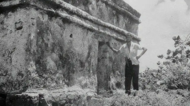 Man standing against weathered stone ruins with vegetation growing around the structure in black and white photograph Man standing against weathered stone ruins with vegetation growing around the structure in black and white photograph
