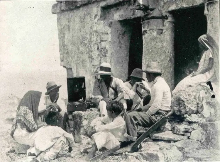 Group of people gathered outside a stone structure with a dark doorway, some wearing hats and traditional clothing in what appears to be a historical photograph Group of people gathered outside a stone structure with a dark doorway, some wearing hats and traditional clothing in what appears to be a historical photograph