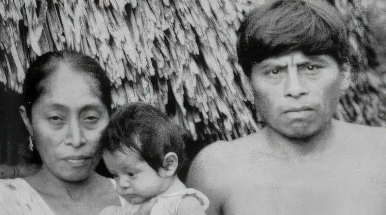 A woman and man stand beside a thatched-roof structure with a young child between them, all looking toward the camera A woman and man stand beside a thatched-roof structure with a young child between them, all looking toward the camera