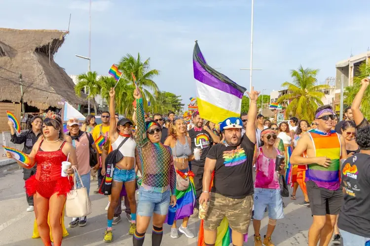 Large crowd of people marching in a street parade while holding rainbow and pride flags under palm trees and clear sky Large crowd of people marching in a street parade while holding rainbow and pride flags under palm trees and clear sky