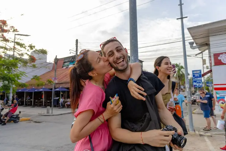 A woman in a pink shirt kisses a man in a black shirt on the cheek while a third person smiles beside them on a busy street lined with shops and utility poles A woman in a pink shirt kisses a man in a black shirt on the cheek while a third person smiles beside them on a busy street lined with shops and utility poles