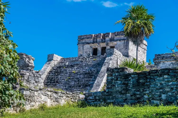 Stone pyramid with watchtower structure stands against blue sky, surrounded by grassy grounds and tropical vegetation
