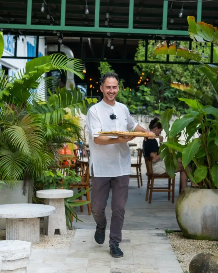 Man in white shirt holding a wooden paddle walks through a plant-filled indoor space with large windows Man in white shirt holding a wooden paddle walks through a plant-filled indoor space with large windows