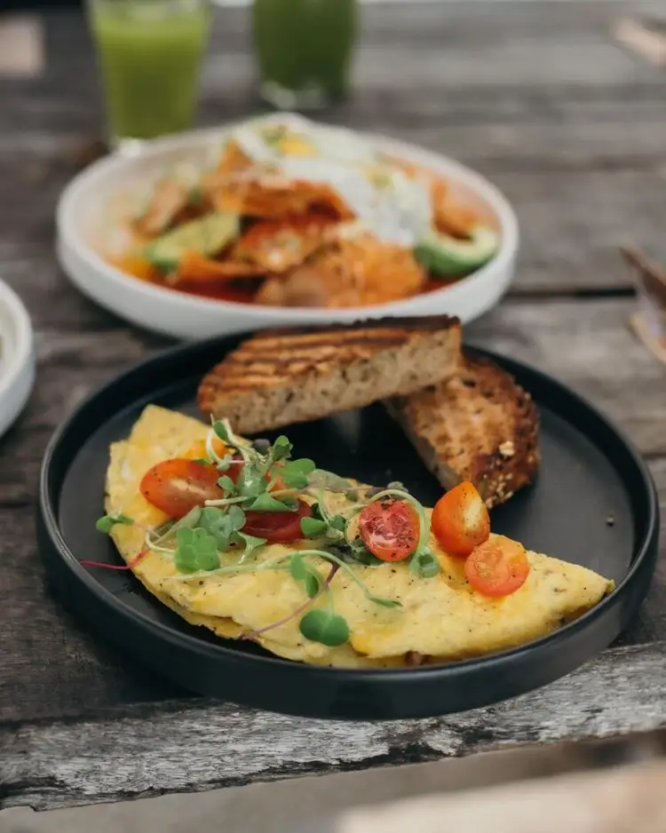 An omelet topped with tomato slices and microgreens sits on a black plate next to toasted bread, with a white plate of vegetables and another beverage in the ba An omelet topped with tomato slices and microgreens sits on a black plate next to toasted bread, with a white plate of vegetables and another beverage in the ba