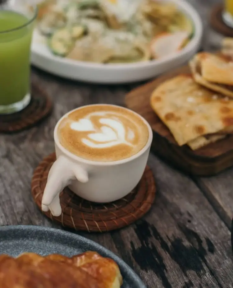 Cappuccino with latte art sits on a wooden coaster surrounded by flatbread, pastries, and a green beverage on a dark table Cappuccino with latte art sits on a wooden coaster surrounded by flatbread, pastries, and a green beverage on a dark table