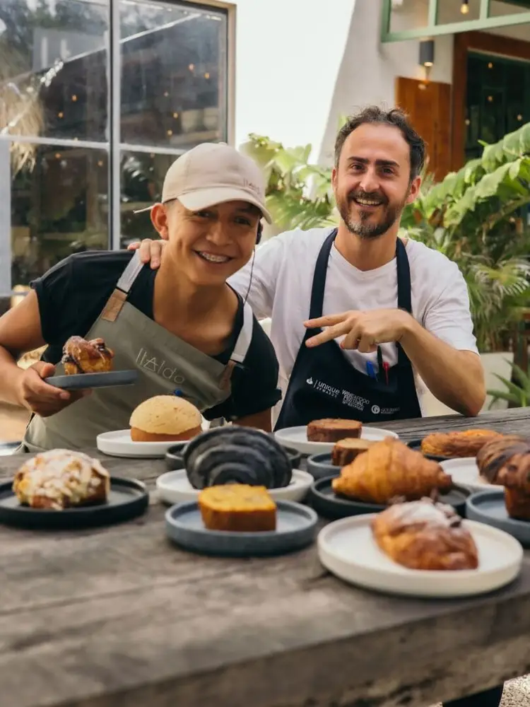 Two people wearing aprons stand behind a table displaying various baked goods including pastries, bread, and cake in a courtyard setting with plants and large w Two people wearing aprons stand behind a table displaying various baked goods including pastries, bread, and cake in a courtyard setting with plants and large w
