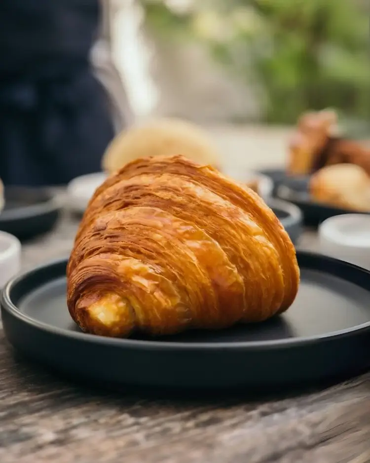 A croissant with golden, flaky layers sits on a dark plate at an outdoor table with other pastries and place settings visible in the background A croissant with golden, flaky layers sits on a dark plate at an outdoor table with other pastries and place settings visible in the background