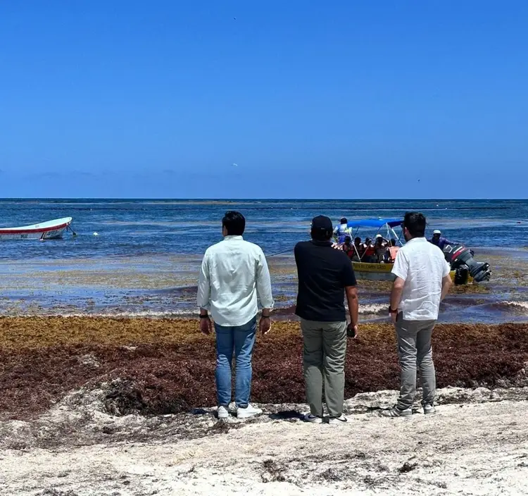 Three men stand on a beach facing the ocean where a boat full of people is visible in shallow water, with sargassum seaweed covering the sandy shore