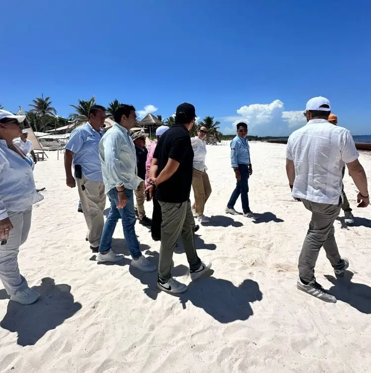 Group of people standing on a sandy beach under clear blue sky, appearing to discuss or inspect the beach area