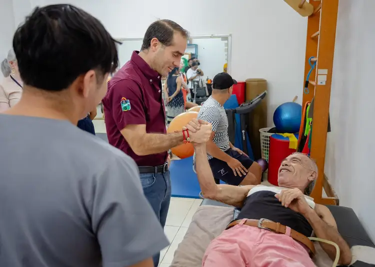 A therapist in a maroon shirt guides an arm exercise with a man lying on a treatment table while another person assists in a therapy room equipped with exercise A therapist in a maroon shirt guides an arm exercise with a man lying on a treatment table while another person assists in a therapy room equipped with exercise