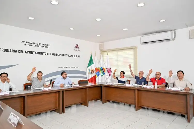 Municipal officials raise their hands during a voting session in a government meeting room with Mexican flags displayed