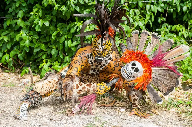 Elaborate ceremonial costume with leopard and jaguar spotted patterns, feathered headdress, and skull mask sits on ground against leafy vegetation