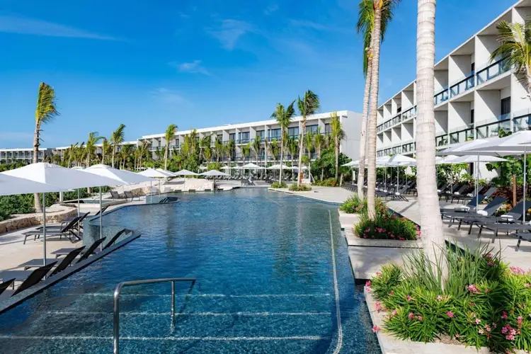 Resort pool surrounded by white umbrellas, lounge chairs, palm trees and multi-story hotel buildings under blue sky Resort pool surrounded by white umbrellas, lounge chairs, palm trees and multi-story hotel buildings under blue sky