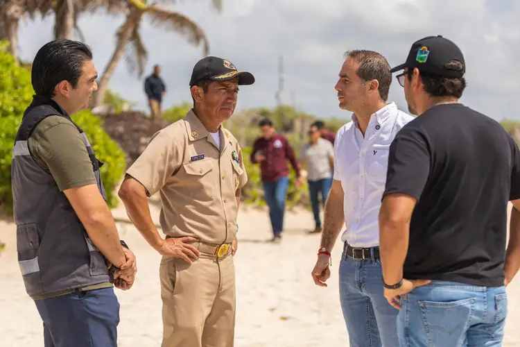 A park ranger in tan uniform speaks with three men on a beach while other visitors stand in the background near palm trees