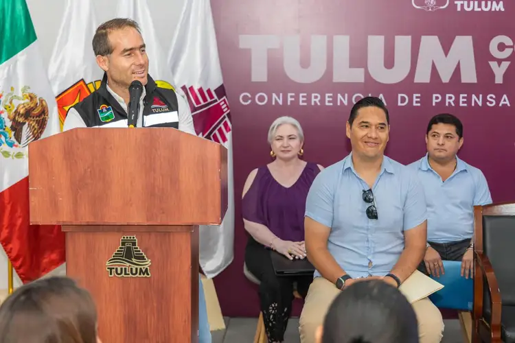 A man in military uniform speaks at a wooden podium while three people sit on a panel behind him during a press conference