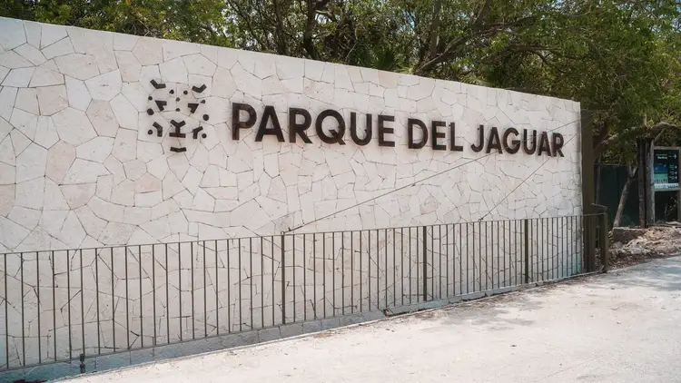 A white stone wall with "Parque del Jaguar" text and logo, fronted by a metal railing with trees visible behind it