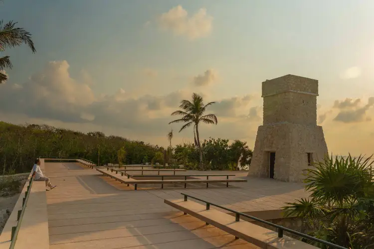 A wooden boardwalk with metal railings leads toward a stone tower structure surrounded by palm trees and tropical vegetation under a cloudy sky