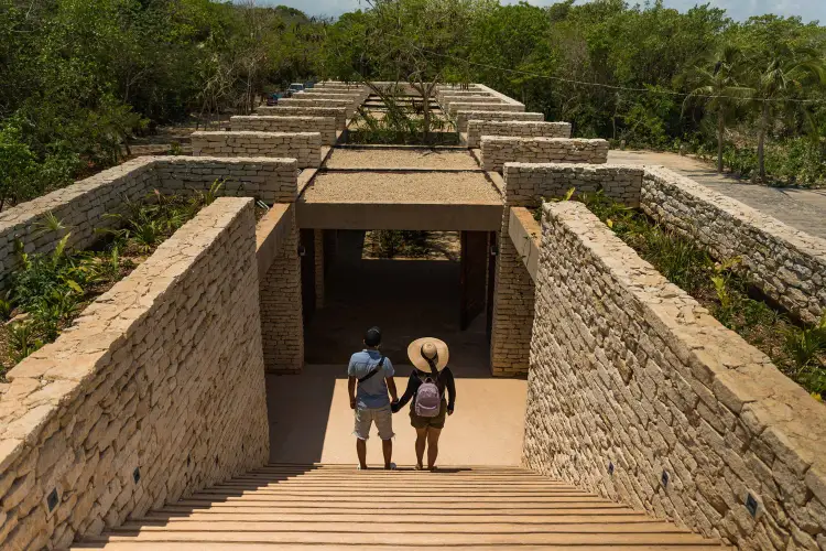 Two visitors walk through a stone passageway leading to a stepped pyramid structure surrounded by dense forest