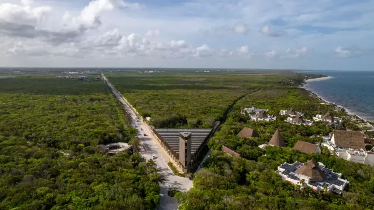Aerial view of a concrete dam structure cutting through dense green forest with a coastal settlement visible on the right and ocean in the distance