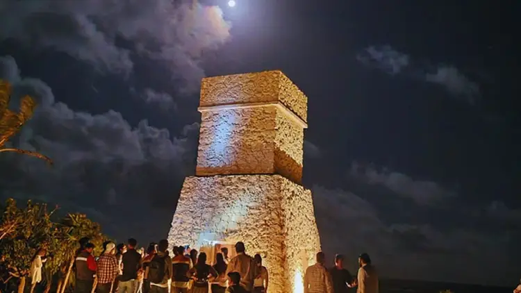 A group of visitors gathers at night around an illuminated stone tower beneath a moon and cloudy sky