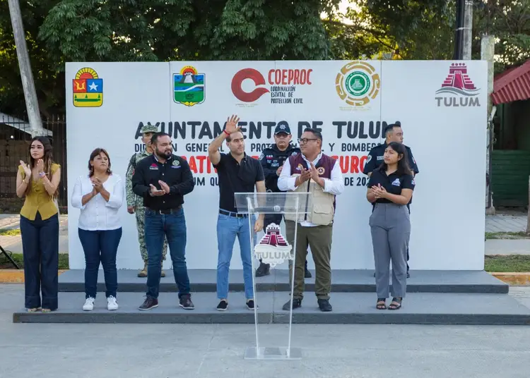A group of officials and representatives stand on a stage with a podium during a public event in Tulum, with municipal and organizational logos displayed on a b A group of officials and representatives stand on a stage with a podium during a public event in Tulum, with municipal and organizational logos displayed on a b