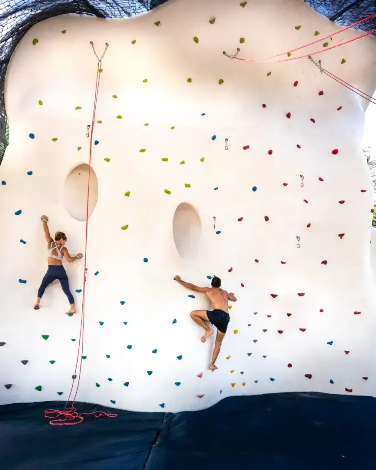 Two climbers scale an inflatable rock wall covered with colorful handholds, one secured by a rope on the left and another climbing freely on the right