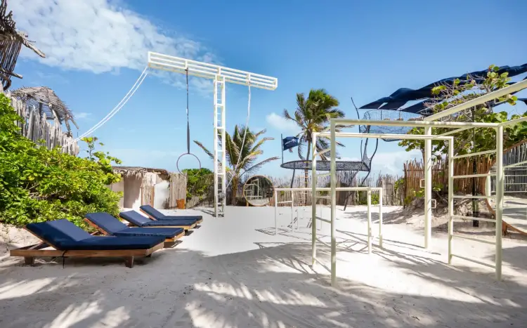Beachfront fitness facility with blue loungers, white pergolas, palm trees, and workout structures on sand under clear sky