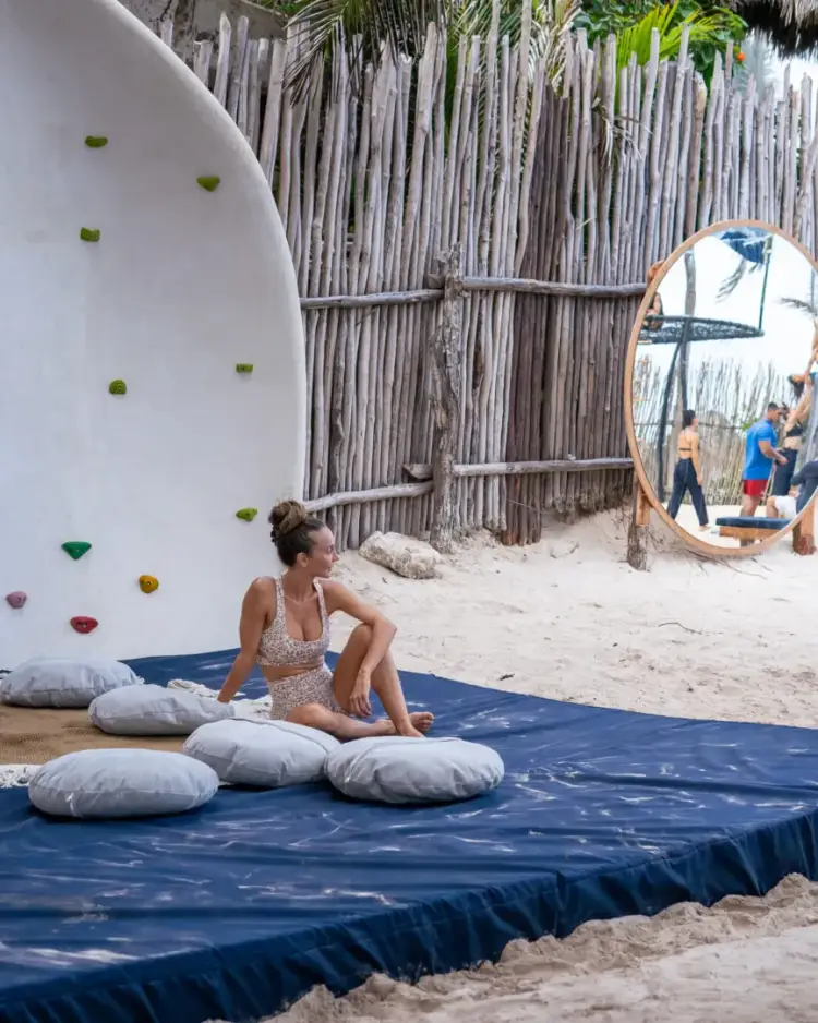 Woman sitting on a blue mat with cushions at a beachfront fitness facility with a climbing wall and mirror nearby