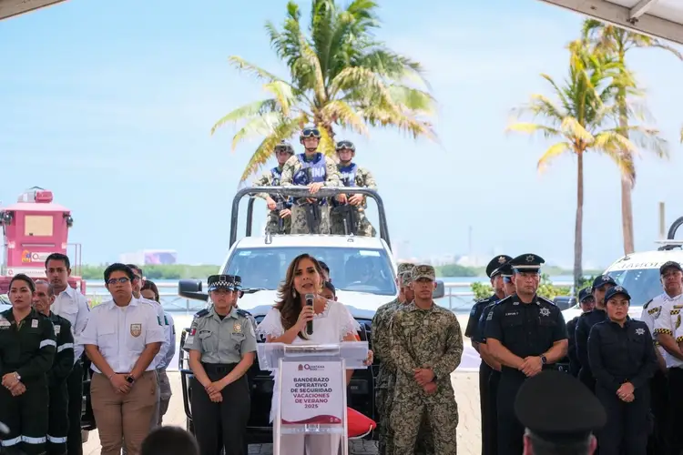Woman speaking at a podium in front of uniformed officials and a vehicle with three people on its roof, with palm trees and clear sky in the background
