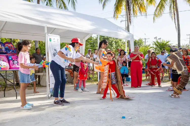 A woman in a red cap leads children in activities under a white tent at an outdoor community gathering with palm trees and attendees in traditional clothing vis