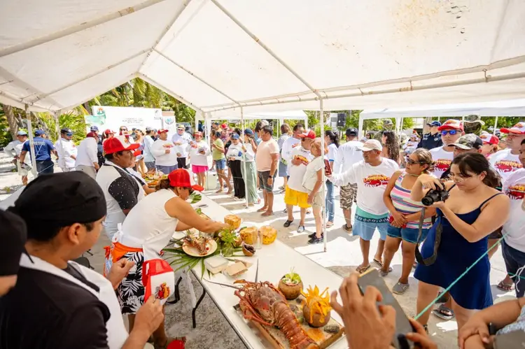 Crowds gather under a tent at an outdoor food festival with vendors serving dishes from tables while attendees sample meals and socialize
