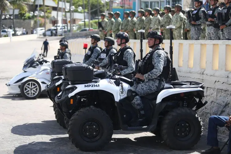 Military personnel on all-terrain vehicles and motorcycles stand in an urban street while uniformed troops gather in the background behind a concrete barrier Military personnel on all-terrain vehicles and motorcycles stand in an urban street while uniformed troops gather in the background behind a concrete barrier